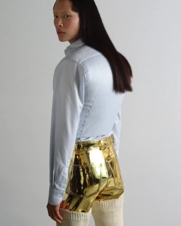 A woman in a light blue button-down shirt and gold shorts poses against a white background, looking over her shoulder.