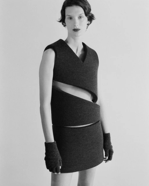 Monochrome studio shot of a woman in a dark gray wool dress and gloves. The dress has layered, spiraling cut-out sections, minimalist style.