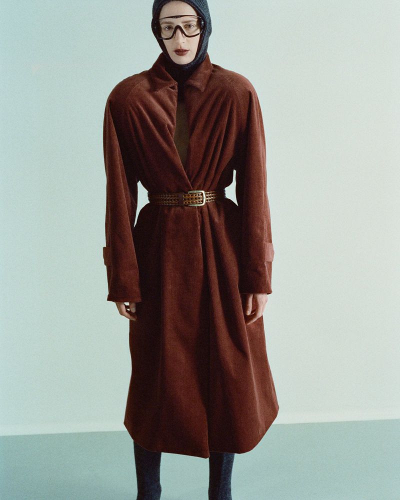 An adult female stands in a light teal studio, wearing a brown corduroy coat, a black knit hood, and clear goggles, creating a unique fashion statement.