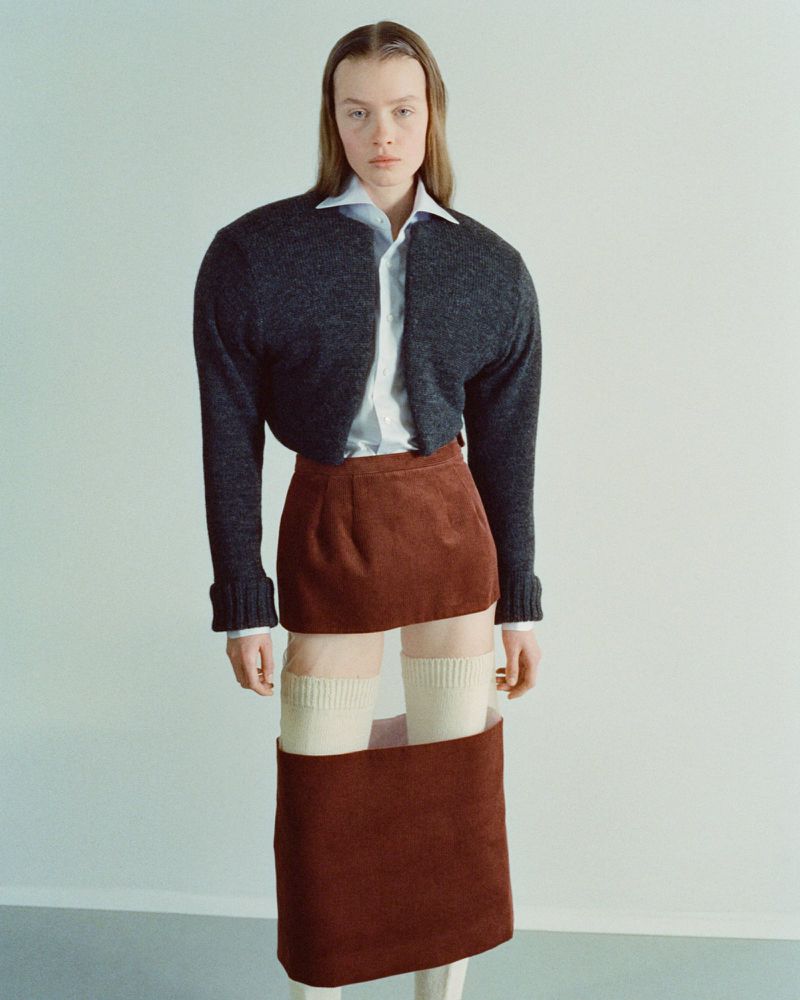 A fashion shot of a fair-skinned woman with straight brown hair posing in a gray cardigan, white button-up shirt, and rust-colored skirt.