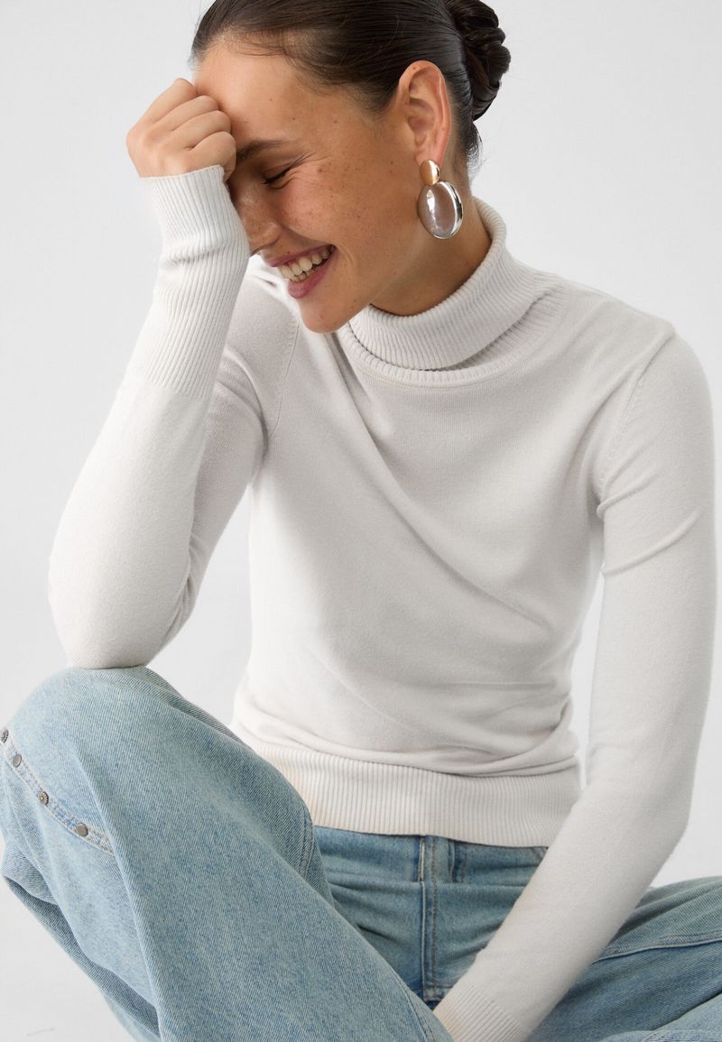 Smiling adult woman in a white turtleneck and jeans, posing indoors with her hand near her face. Statement earrings are visible.