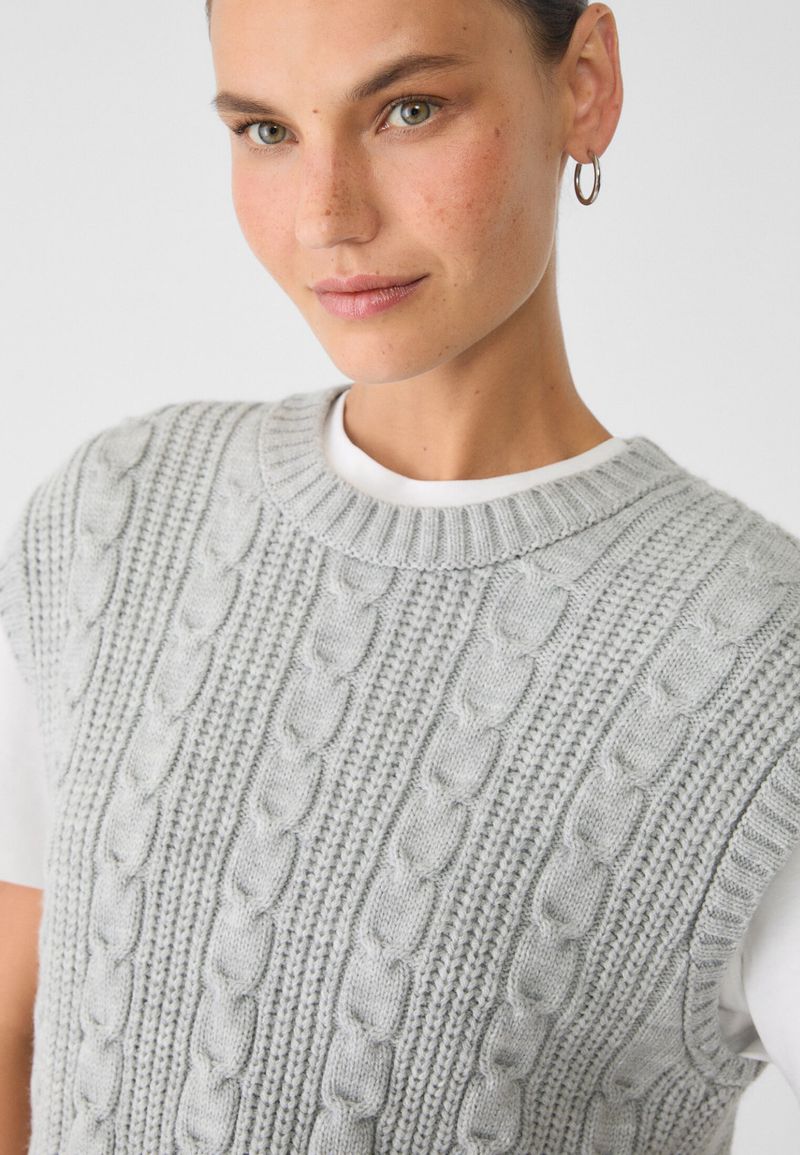 Adult female wearing a gray knit sweater vest over a white t-shirt, hoop earrings, photographed in a close-up studio shot.