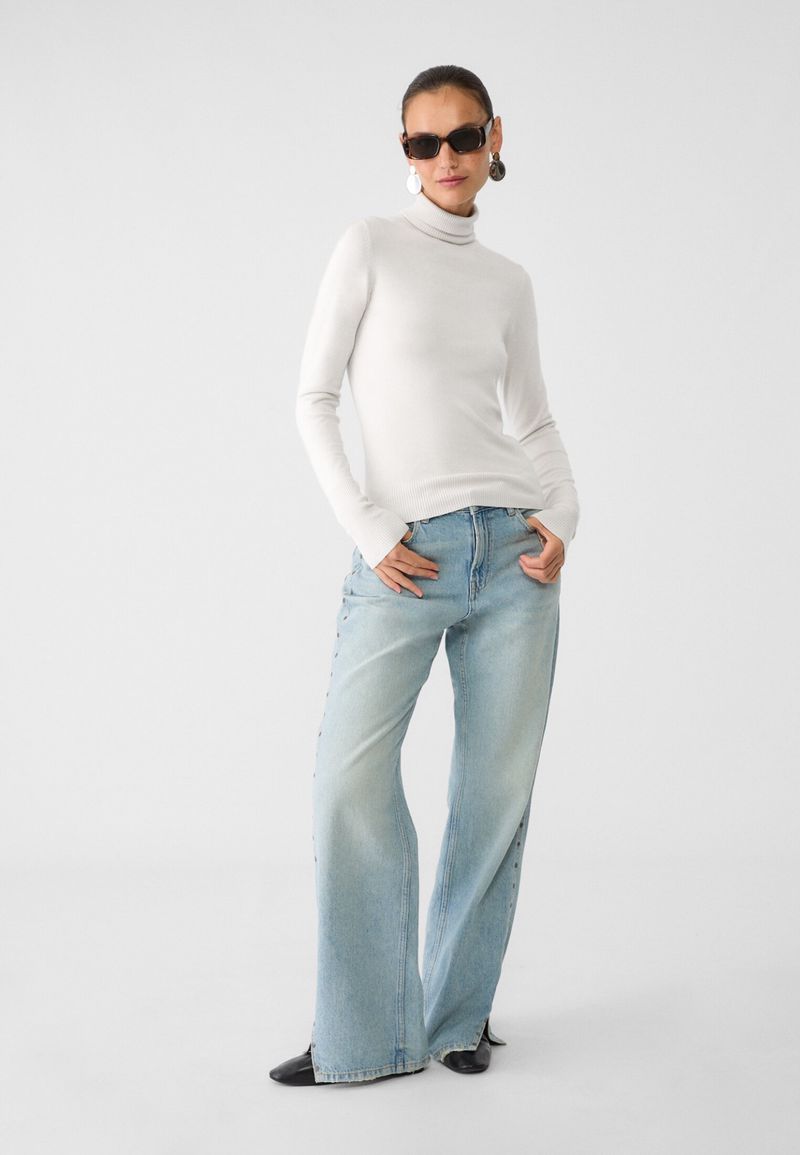 Fashion model poses in a long-sleeve top and wide-leg jeans, accessorized with sunglasses and earrings, against a plain white backdrop.