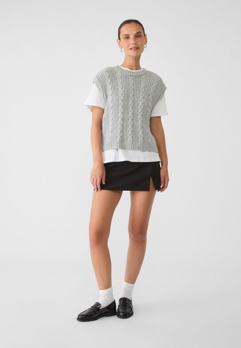 Studio shot of a woman in a knitted vest, mini skirt, and loafers. Full-length portrait against a white backdrop, fashion editorial look.