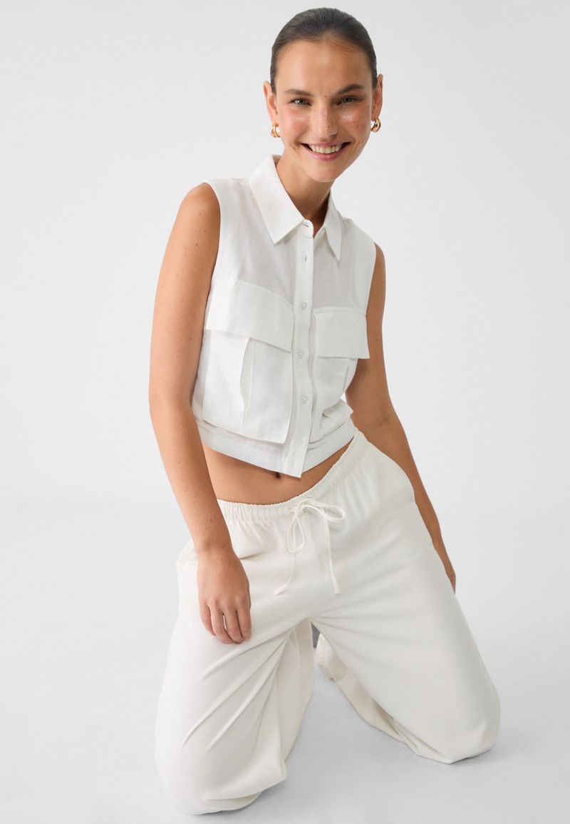 Woman posing in a sleeveless button-down top and loose white pants in a studio environment with a clean white backdrop and soft lighting.