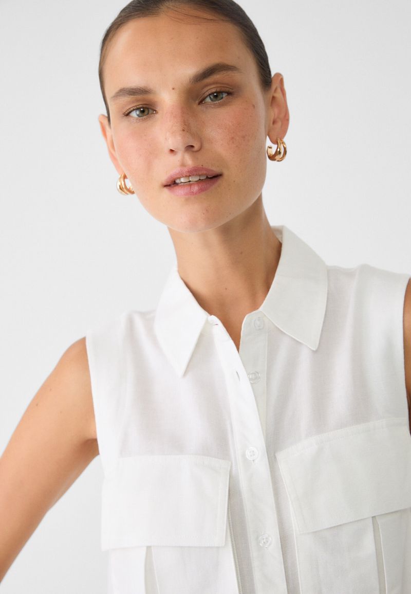 Close-up studio shot of a fair-skinned adult female wearing gold hoop earrings and a sleeveless collared white cotton blouse.