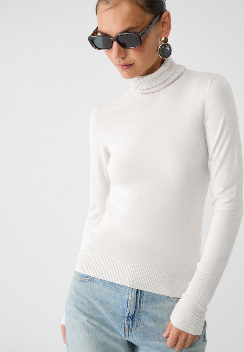 Woman in a white turtleneck sweater and blue jeans wearing sunglasses and circular earrings against a plain white background.