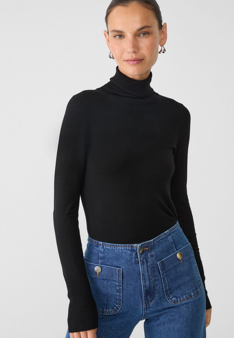A woman poses in a black turtleneck sweater and blue jeans with front pockets, in a medium studio shot with soft lighting.