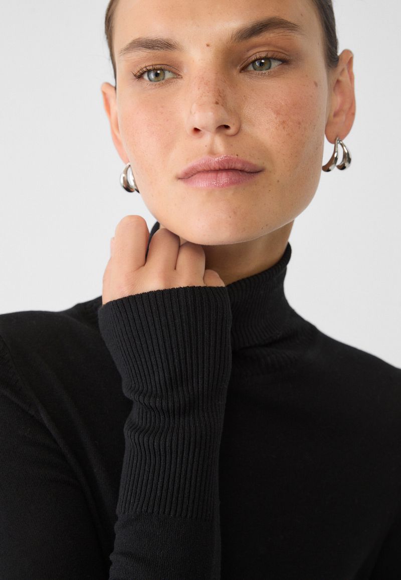 Close-up portrait of a woman in a black ribbed turtleneck sweater and silver drop earrings against a white background.