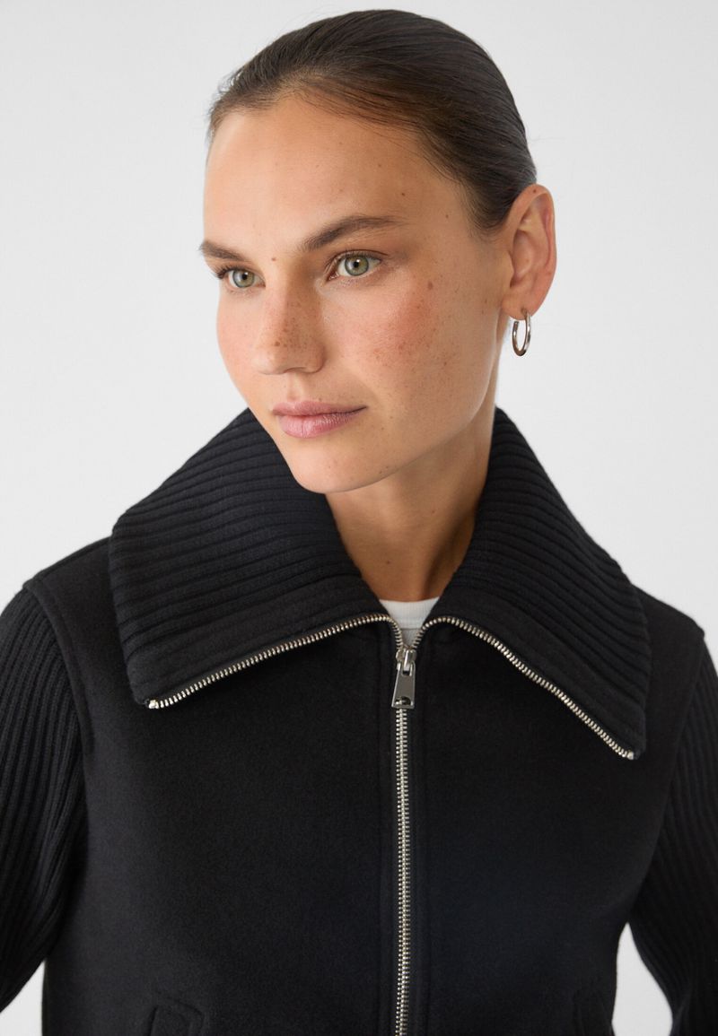Close-up portrait of a woman with hoop earrings wearing a black zip-up jacket with a ribbed collar against a neutral background.