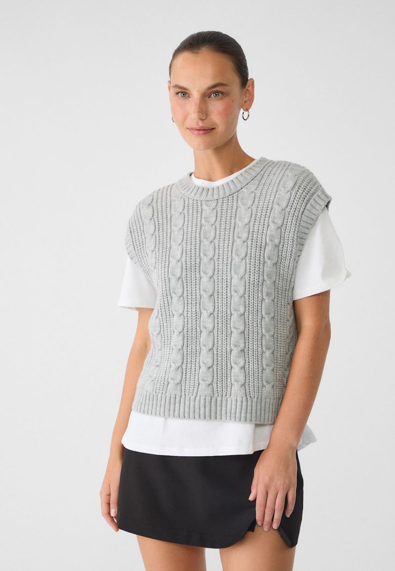 A woman is posing indoors, wearing a gray knit sweater vest over a white t-shirt and a black skirt in a clean, neutral setting.