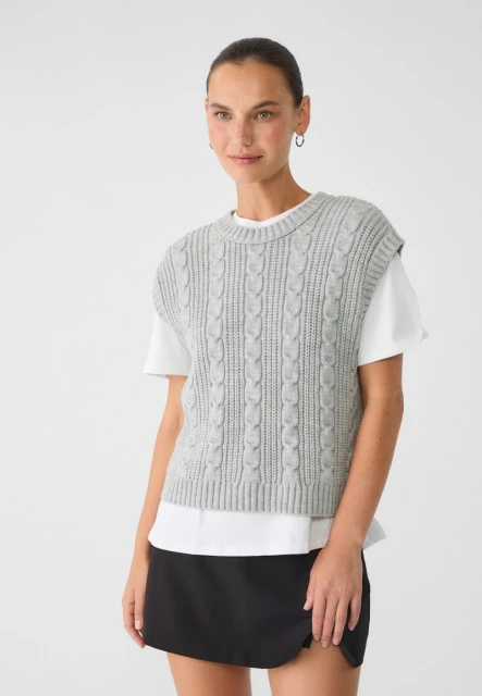 A woman is posing indoors, wearing a gray knit sweater vest over a white t-shirt and a black skirt in a clean, neutral setting.