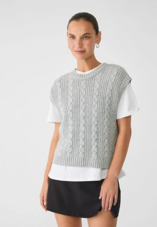 A woman is posing indoors, wearing a gray knit sweater vest over a white t-shirt and a black skirt in a clean, neutral setting.