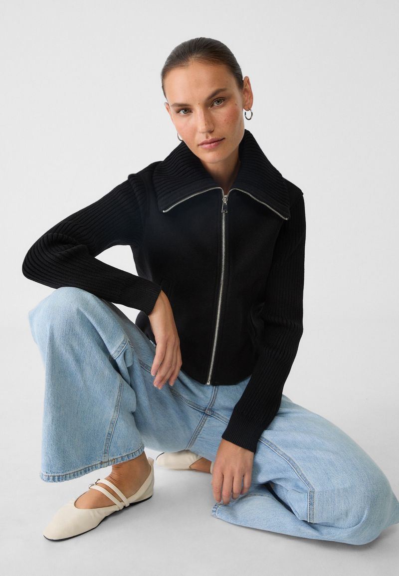 Woman wearing a black zip-up sweater and blue jeans, posing in a photo studio with a bright white background. Fashion lifestyle.