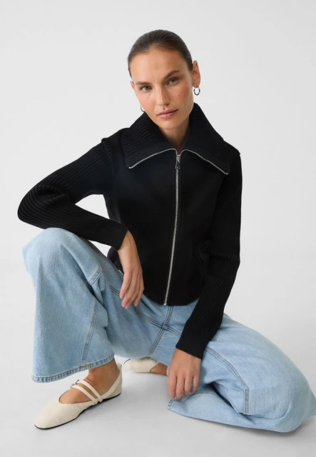 Woman wearing a black zip-up sweater and blue jeans, posing in a photo studio with a bright white background. Fashion lifestyle.