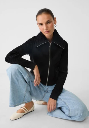 Woman wearing a black zip-up sweater and blue jeans, posing in a photo studio with a bright white background. Fashion lifestyle.