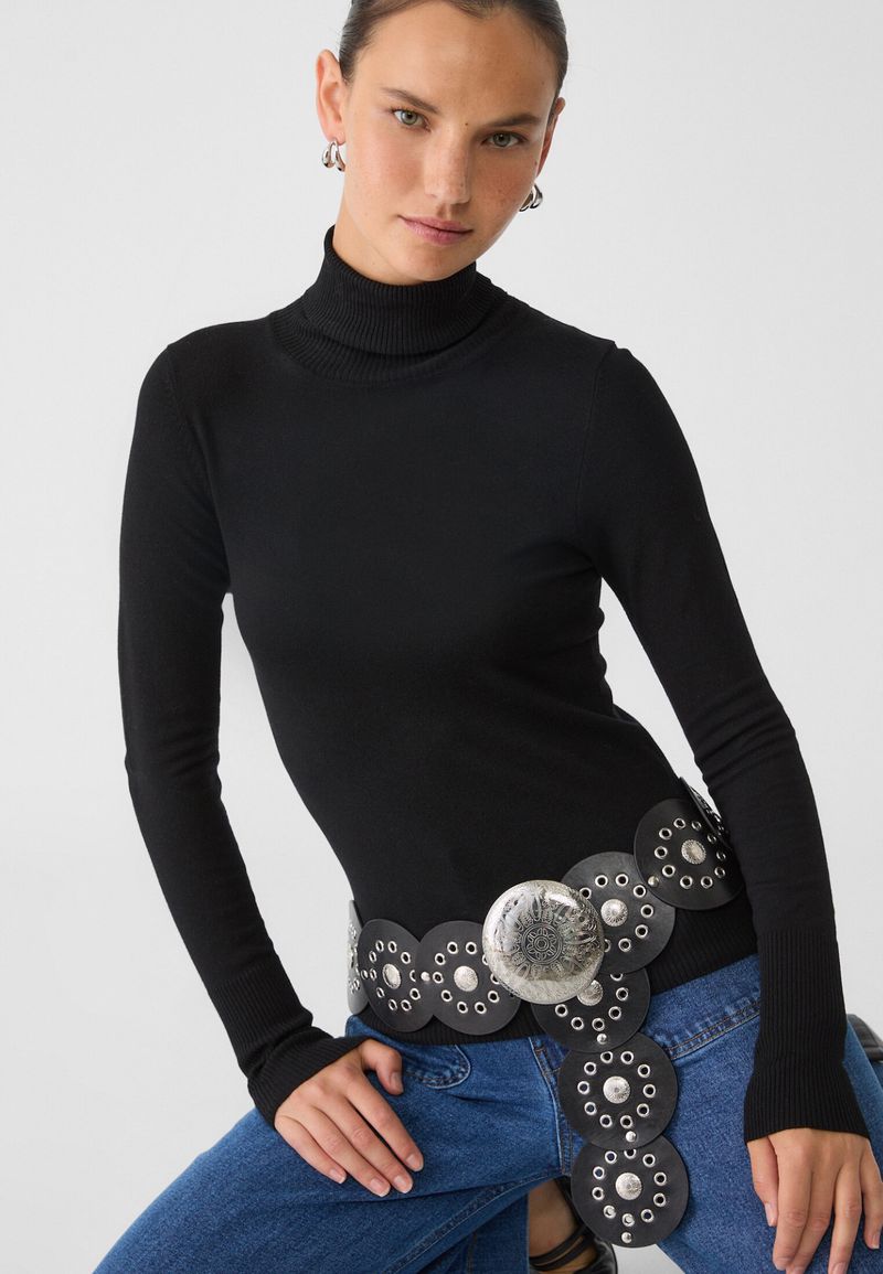 Studio shot of a woman in a black turtleneck, blue jeans, and a studded belt. The image focuses on fashion and stylish casual wear.