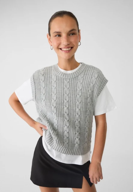 Smiling woman in a gray cable-knit vest over a white t-shirt and black skirt poses in front of a neutral studio backdrop.