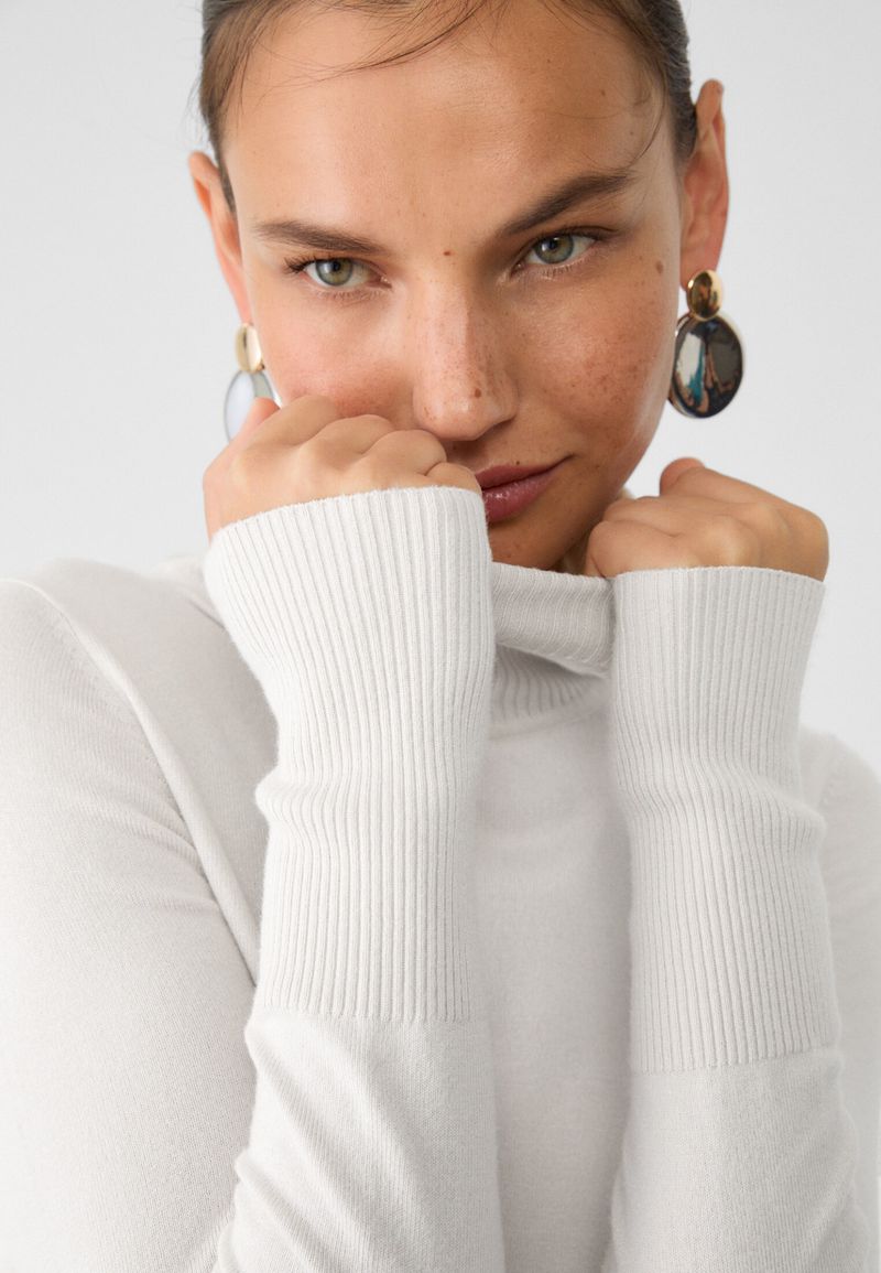 Close-up studio portrait of an adult woman with light skin, wearing a white turtleneck sweater and gold earrings, hands near her face.