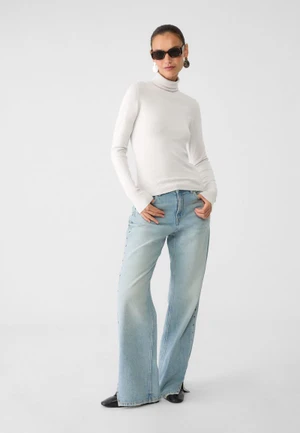 Model wearing a white turtleneck sweater, wide-leg light blue jeans, and dark sunglasses against a seamless white studio background.