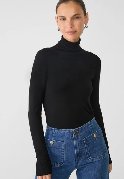 Model wearing a black turtleneck sweater and blue denim jeans against a plain white studio background.