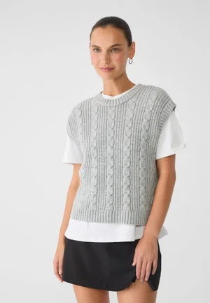Model wearing a grey cable knit sweater vest over a white t shirt and black skirt against a plain white background