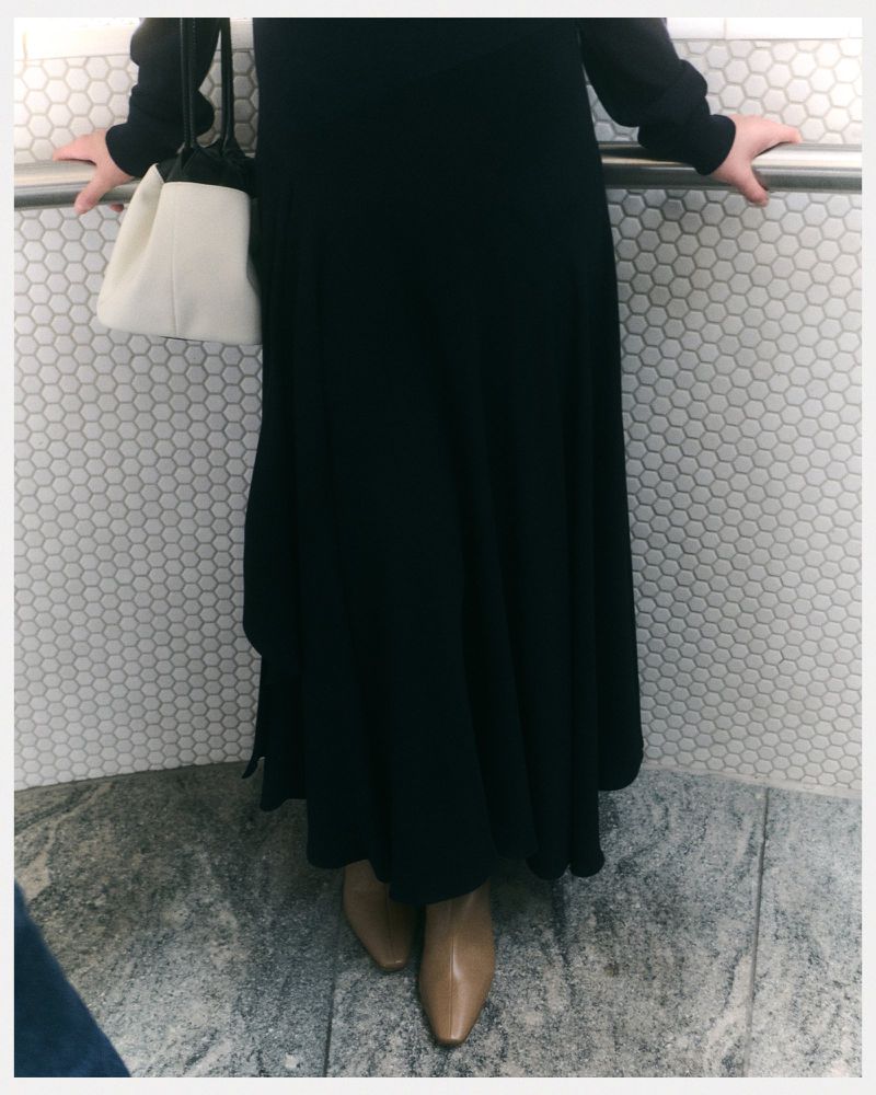 A woman in a black dress stands against a white tile wall with a gray floor, holding a light-colored purse and wearing tan ankle boots.