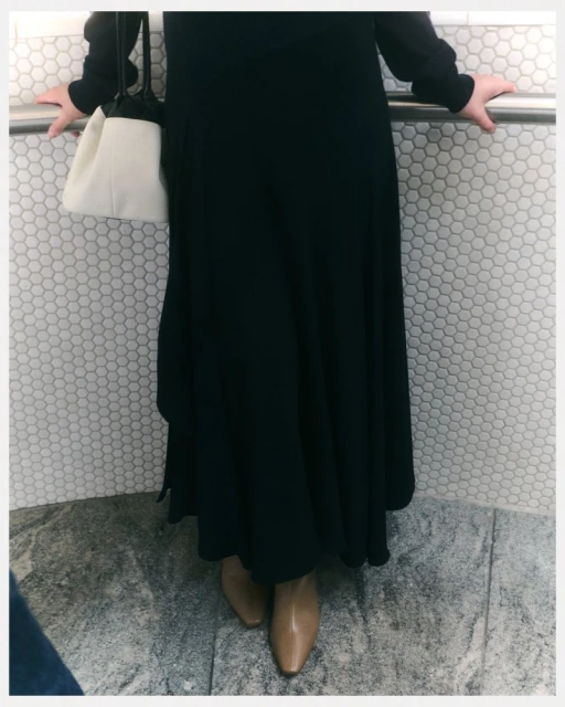 A woman in a black dress stands against a white tile wall with a gray floor, holding a light-colored purse and wearing tan ankle boots.
