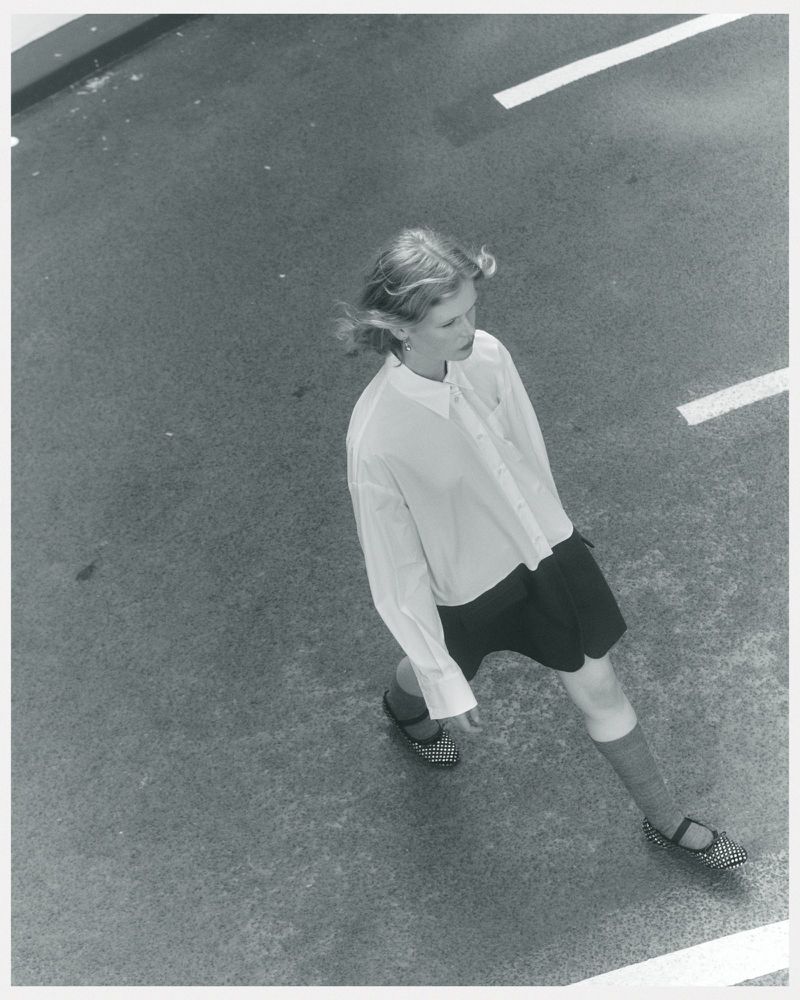 Overhead shot of a fair-skinned child in a white blouse and dark shorts, walking across a gray surface marked with white lines.