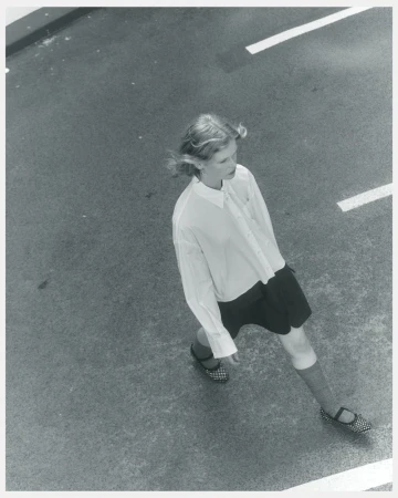 Overhead shot of a fair-skinned child in a white blouse and dark shorts, walking across a gray surface marked with white lines.