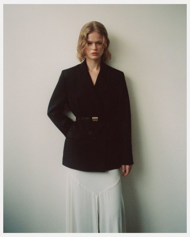 A fashionable blonde woman stands indoors against a cream wall wearing a black, belted blazer and a long white skirt, looking ahead.