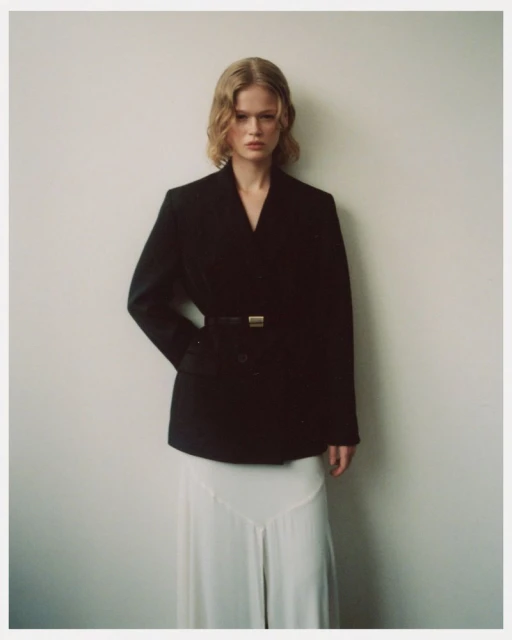 A fashionable blonde woman stands indoors against a cream wall wearing a black, belted blazer and a long white skirt, looking ahead.