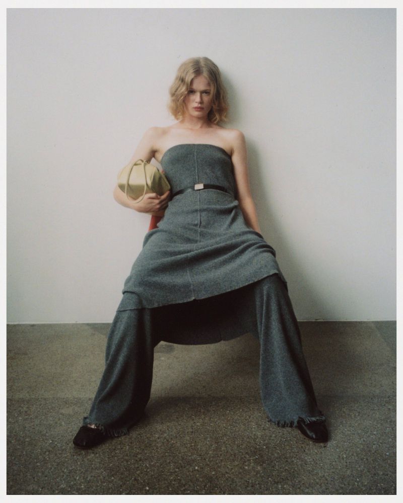 Fashion shot of a woman with blonde hair sitting on a red chair in a strapless gray dress, holding a gold handbag against a white wall.