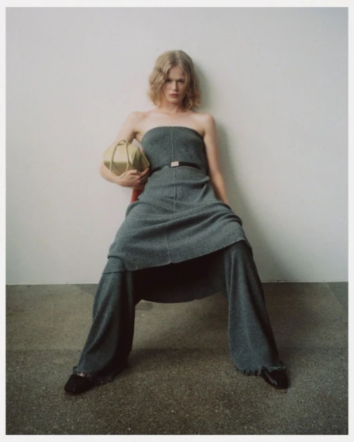 Fashion shot of a woman with blonde hair sitting on a red chair in a strapless gray dress, holding a gold handbag against a white wall.