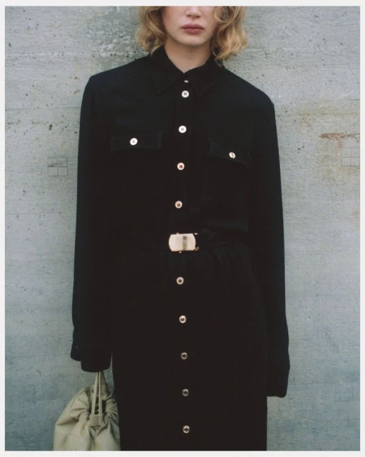 An adult woman poses in a black collared button-down dress with a silver buckle belt and a soft white bag in a studio setting.