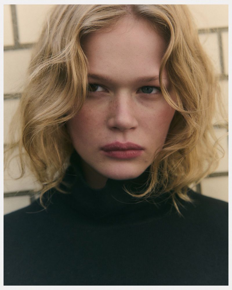 Close-up portrait of a blonde woman with fair skin wearing a black turtleneck against a neutral tile background with soft, diffused lighting.