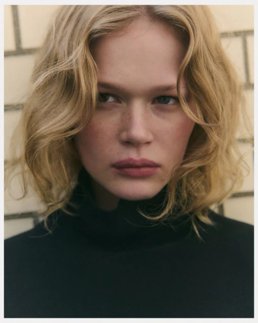 Close-up portrait of a blonde woman with fair skin wearing a black turtleneck against a neutral tile background with soft, diffused lighting.