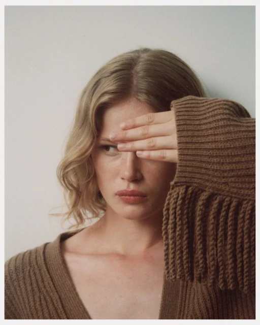 Blonde woman with hand covering eye, portrait in a brown knit cardigan against a white backdrop in soft, natural light.