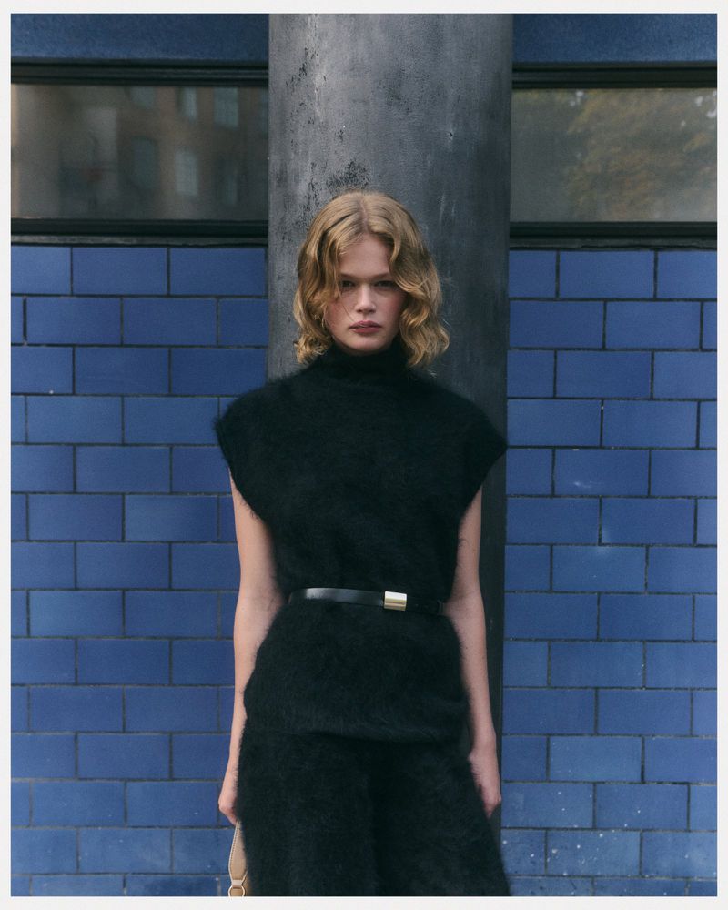 Blonde model poses in a black mohair sweater and pants, cinched at the waist with a belt, against a blue tiled wall indoors.