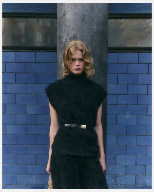 Blonde model poses in a black mohair sweater and pants, cinched at the waist with a belt, against a blue tiled wall indoors.