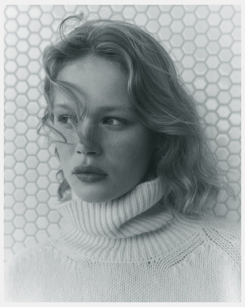Portrait of a fair-skinned woman with wavy hair, wearing a turtleneck sweater, posing in front of a hexagonal tile wall.