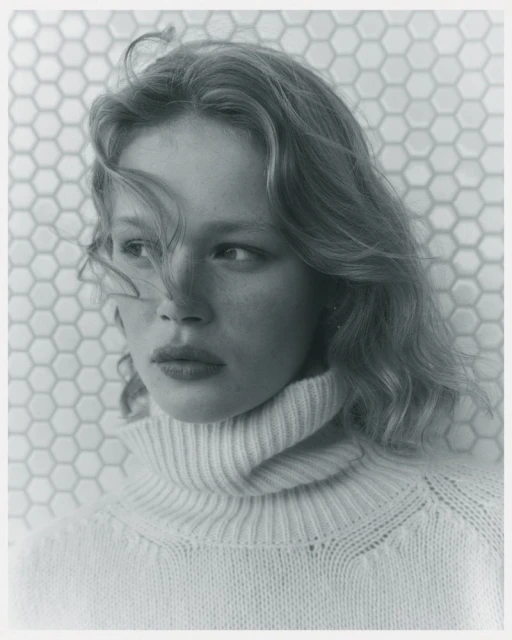 Portrait of a fair-skinned woman with wavy hair, wearing a turtleneck sweater, posing in front of a hexagonal tile wall.