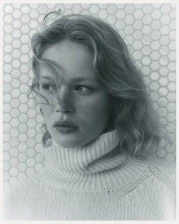 Portrait of a fair-skinned woman with wavy hair, wearing a turtleneck sweater, posing in front of a hexagonal tile wall.