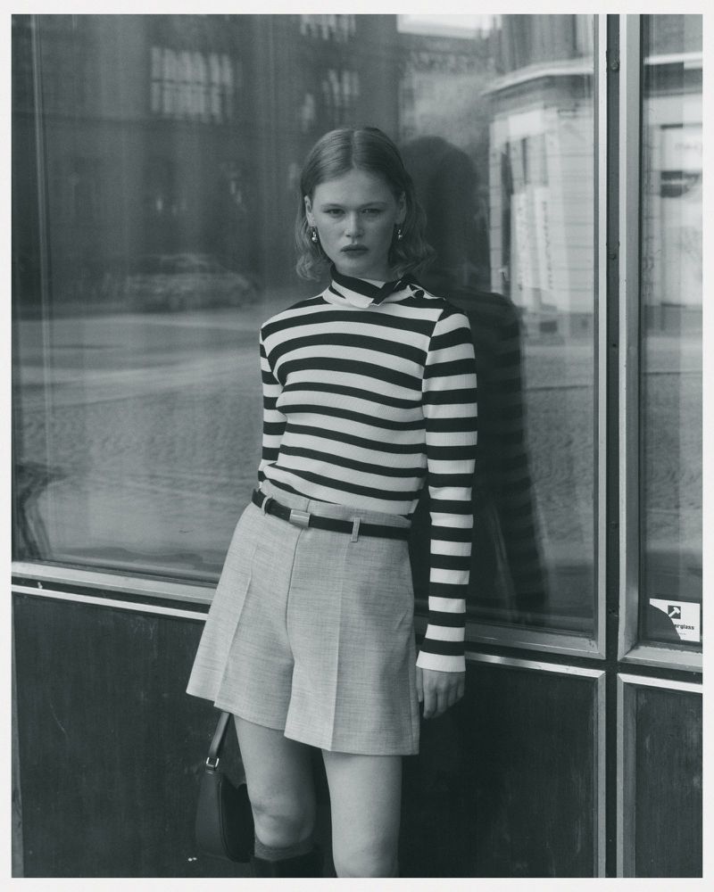 A fair-skinned woman with curly hair poses outdoors in a striped long-sleeve shirt, grey shorts, black boots, and a small black purse.