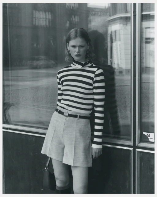 A fair-skinned woman with curly hair poses outdoors in a striped long-sleeve shirt, grey shorts, black boots, and a small black purse.