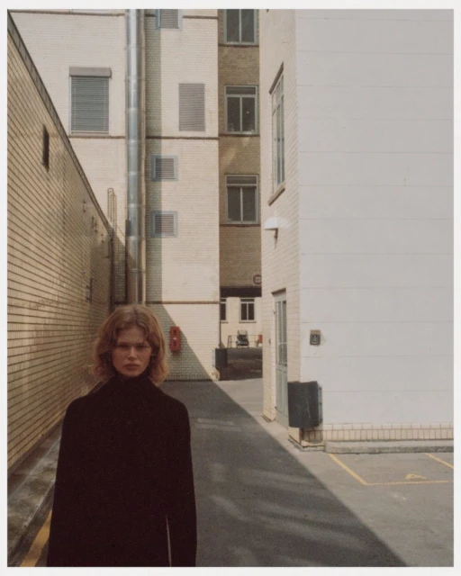 Portrait of an adult woman with curly auburn hair, wearing a black top, standing in an alleyway with brick and concrete buildings.
