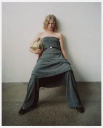 Model wearing a grey strapless dress and trousers holding a light yellow handbag against a white wall.
