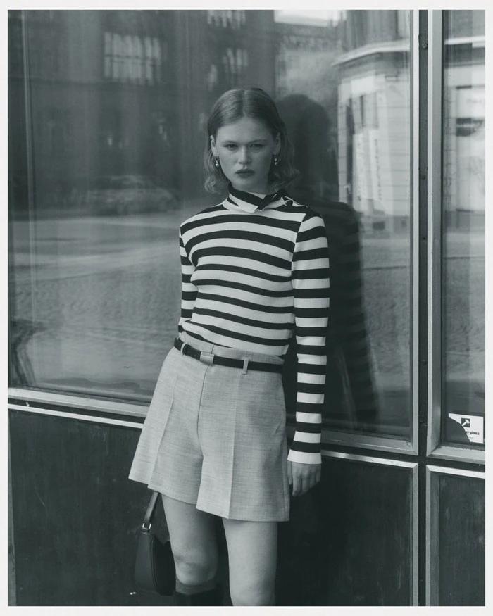 Model wearing a black and white striped top and shorts leaning against a glass storefront in a monochrome fashion portrait