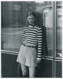 Model wearing a black and white striped top and shorts leaning against a glass storefront in a monochrome fashion portrait