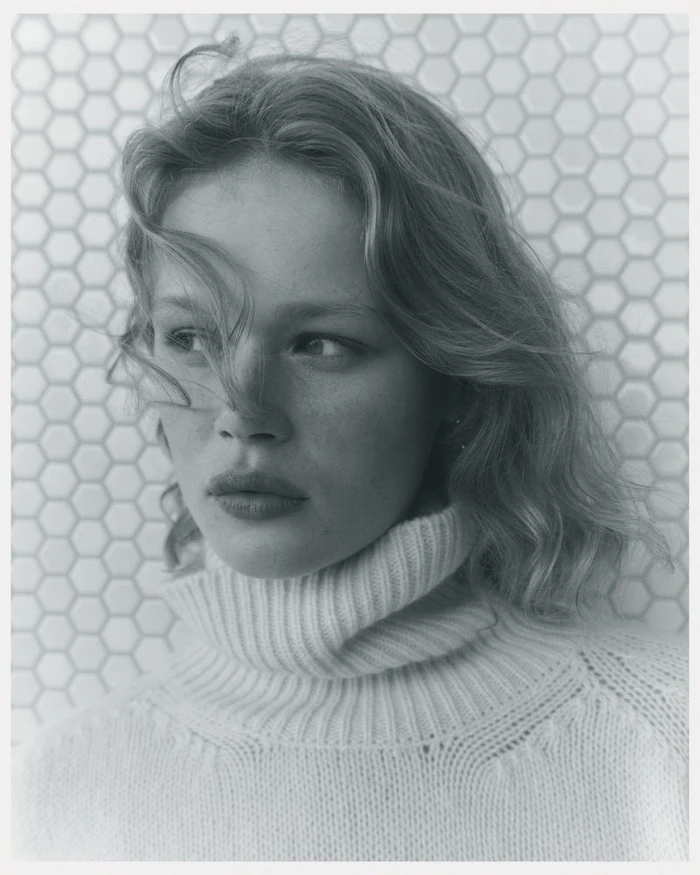 Portrait of a female model wearing a cream turtleneck sweater against a patterned tiled background.